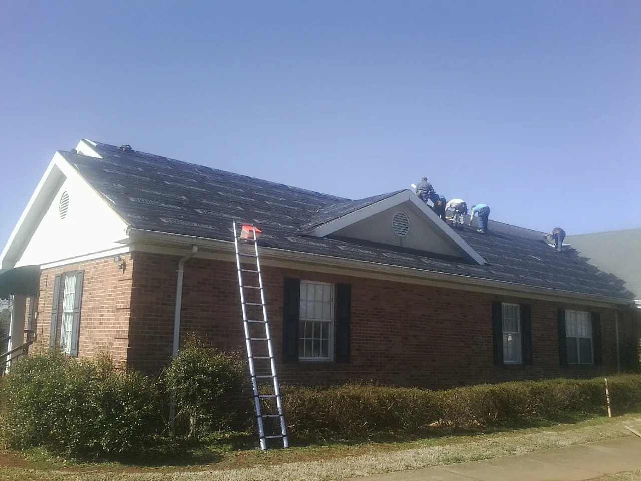 Crew tearing shingles off a brick ranch roof with a ladder set — Emergency Roof Repair work by our crew in Ballston Spa