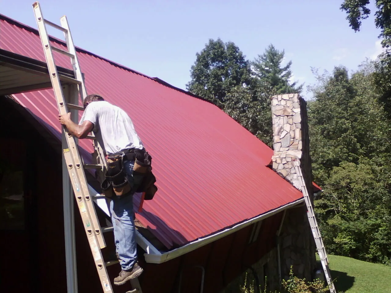 Roofer on a ladder beside a red metal roof and stone chimney — Soffit & Fascia Repair by Roofing Brothers in Ballston Spa